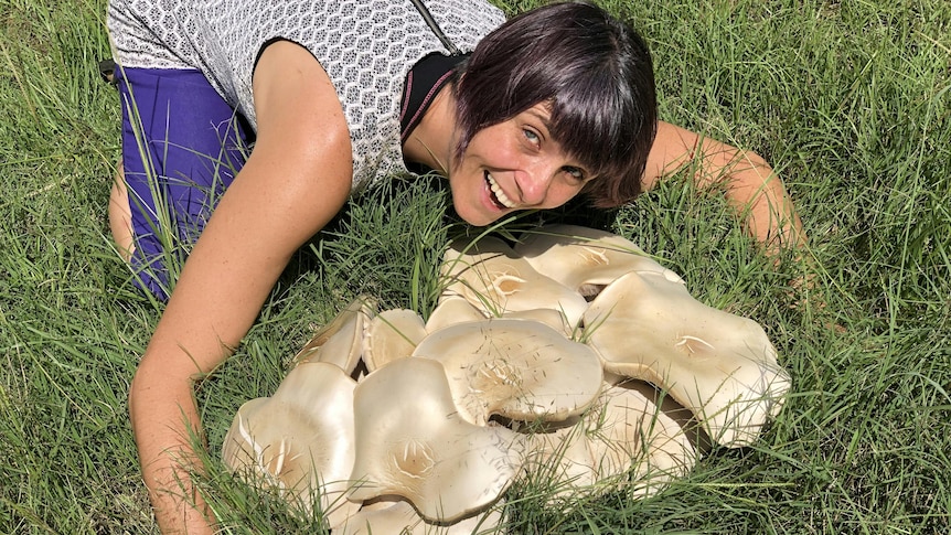 A middle-aged woman with a brown bob haircut smiles as she hugs a large mushroom formation in a field.