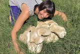 A middle-aged woman with a brown bob haircut smiles as she hugs a large mushroom formation in a field.