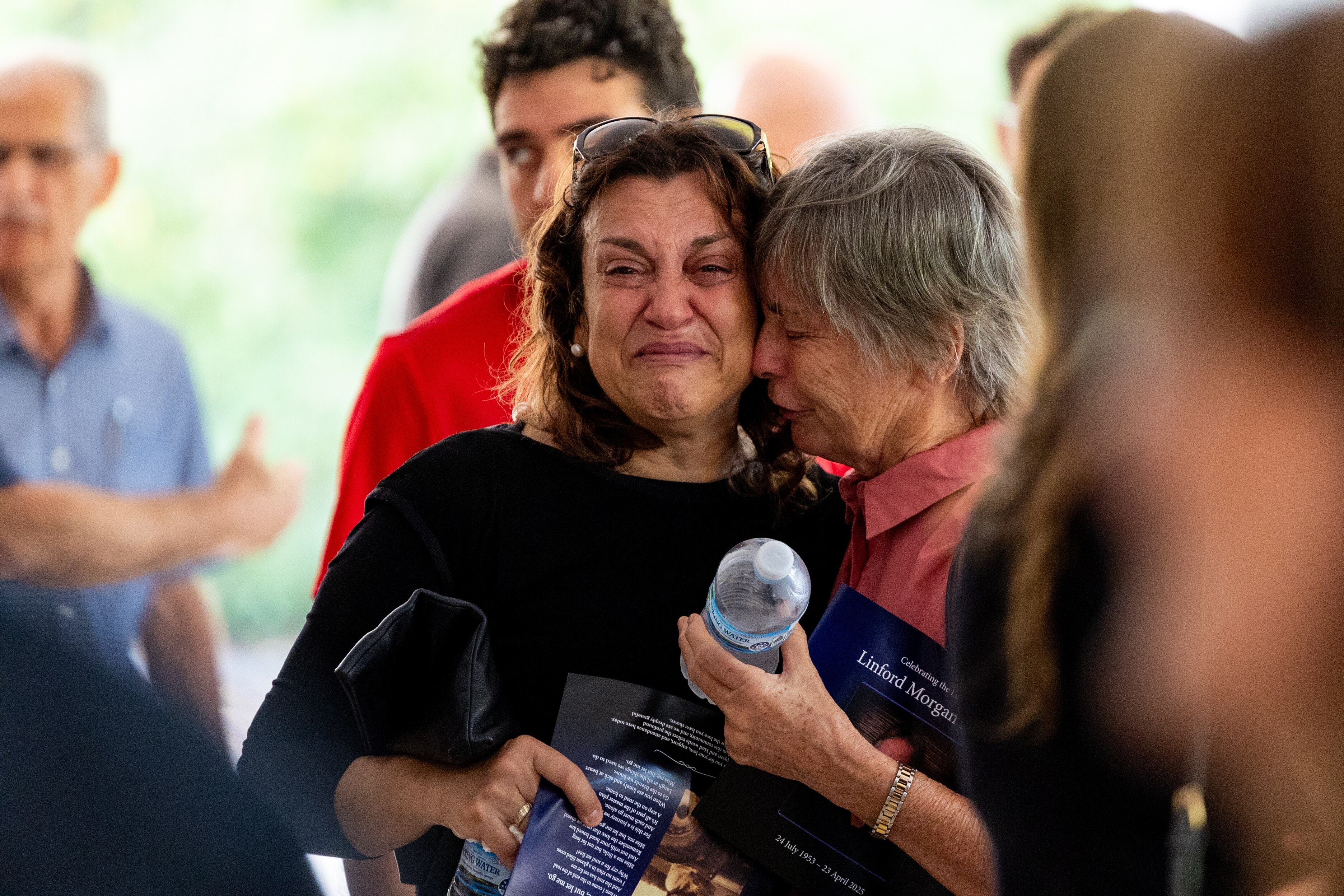 Two women in focus. One wearing black, flushed face and crying. Another woman in pink shirt is comforting her.