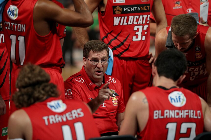 The Perth Wildcats NBL coach crouches as he speaks to his player who are surrounding him.