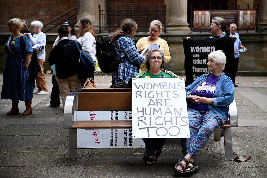 A group of people sit outside a court holding placards and signs.