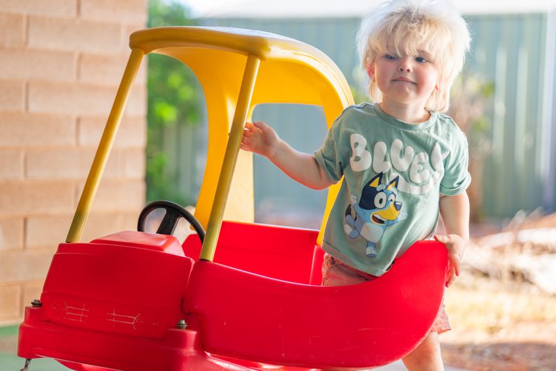 Un niño pequeño sonriente con una camisa azul se encuentra junto a un coche de juguete con pedales rojo y amarillo. 