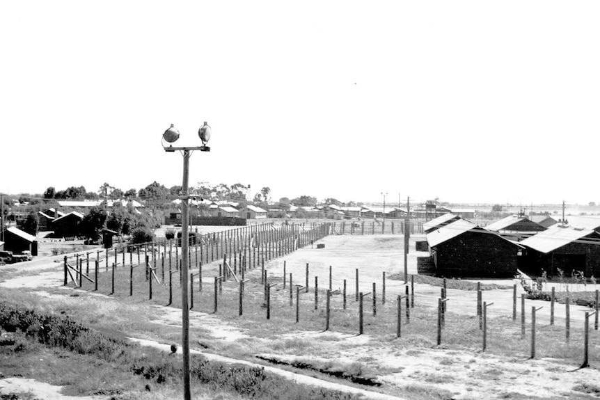 Black and white photo of camp buildings surrounded by several lines of barbed wire fence.