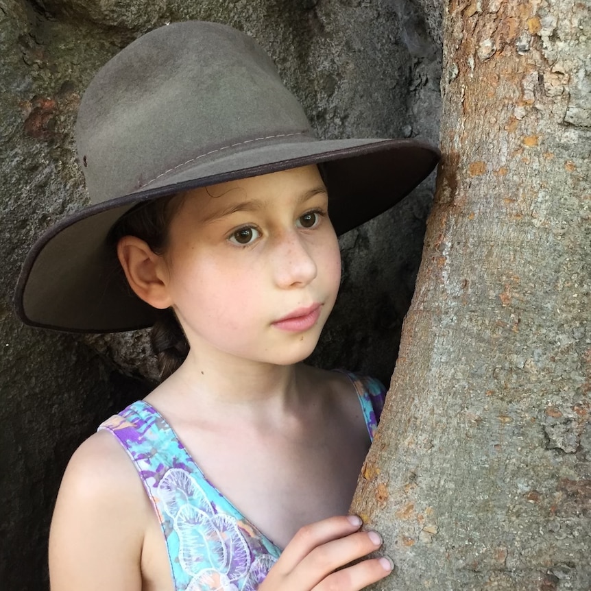 A young girl wearing a wide-brimmed hat stands next to the trunk of a tree.