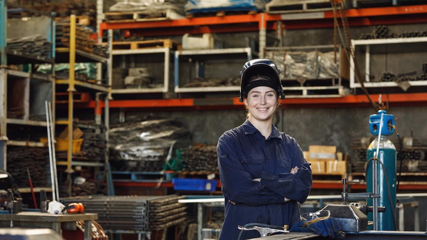 A young woman stands in a workshop in blue overalls wearing a raised welding visor. She is smiling and her arms are crossed.