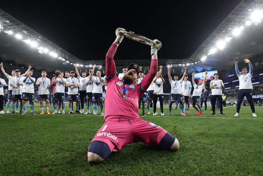 A goalkeeper in a pink jersey and shorts leans back as he kneels on the grass, raising a trophy while his teammates cheer.