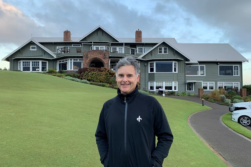 A man in a warm jacket stands in front of a green golf course and clubhouse.
