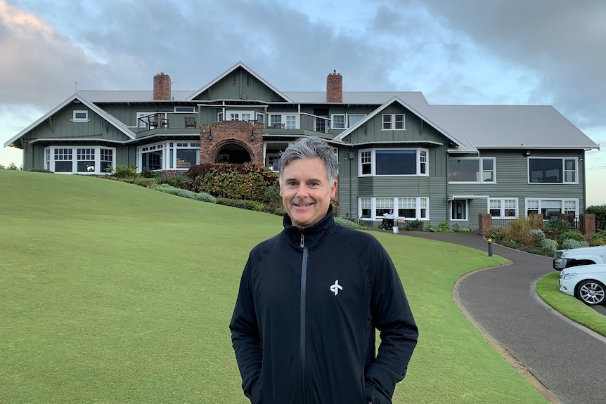 A man in a warm jacket stands in front of a green golf course and clubhouse.