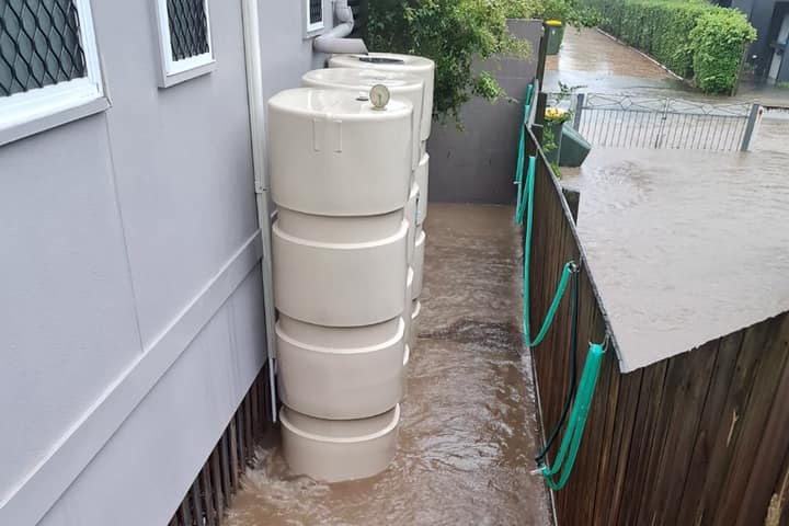 Brown storm water flowing under a high-set house and pooling between a beige water tank and wooden fence.