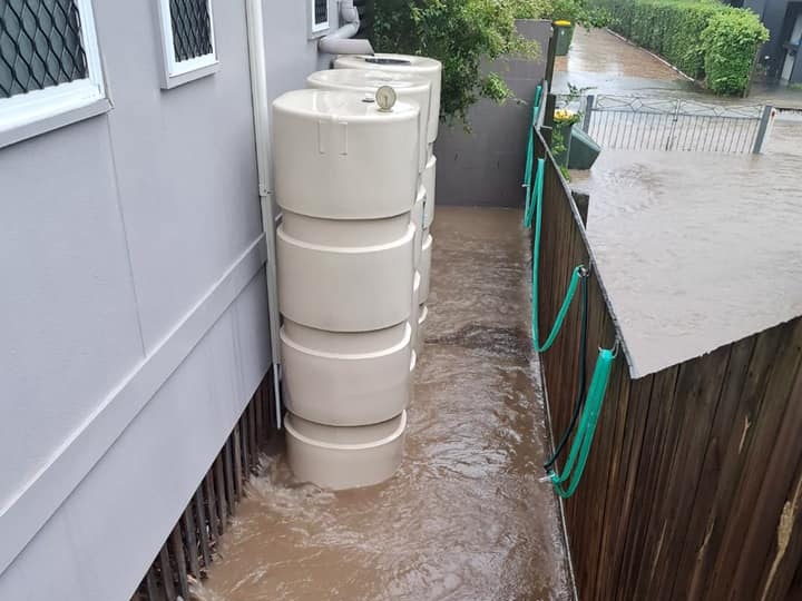 Brown storm water flowing under a high-set house and pooling between a beige water tank and wooden fence.