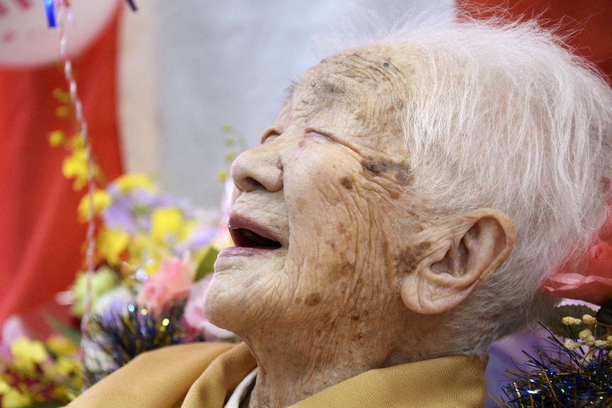 An elderly Japanese woman smiles with an open mouth and closed eyes, with flowers behind her head.