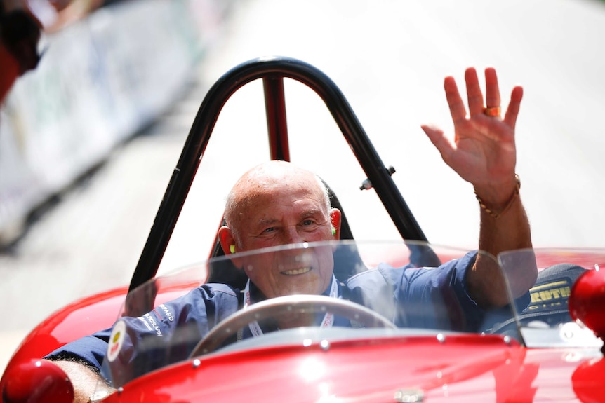 Stirling Moss, sitting in a red F1 car, waves to a crowd.