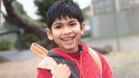 A five-year-old boy holds his backpack and smiles.