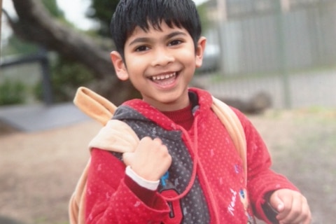 A five-year-old boy holds his backpack and smiles.