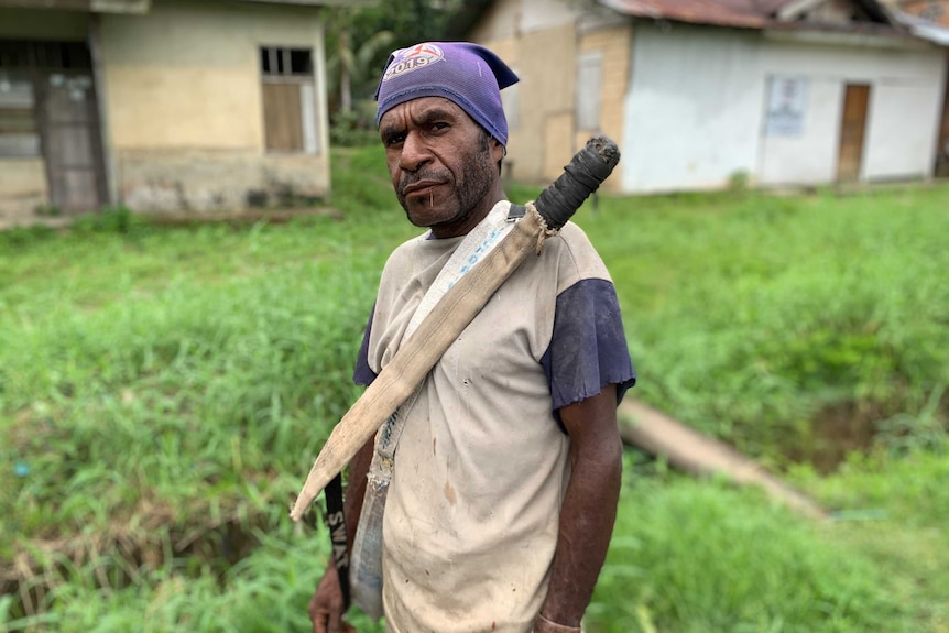 A man stands in a field. A machete in a sheath is attached to his chest.