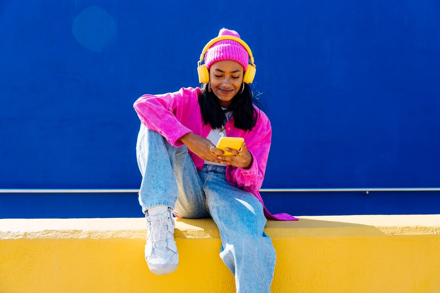 A young woman with dark skin and black hair wearing yellow headphones looks at a yellow smartphone 