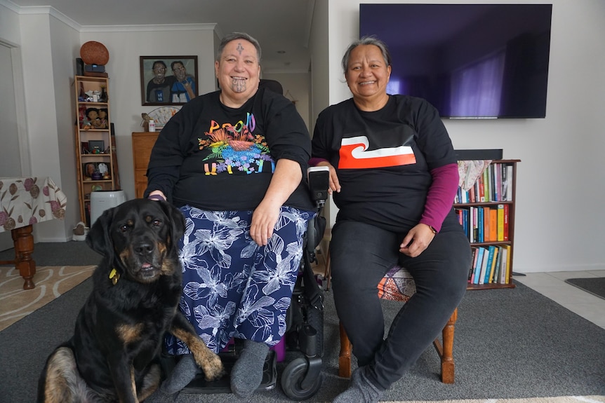 Huhana is wearing a black printed tee with blue and white colourful pants  sitting next to her partner and service dog