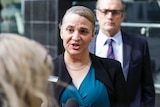 A woman in a blue dress and black jacket speaks to a journalist with a man in a suit standing behind her.