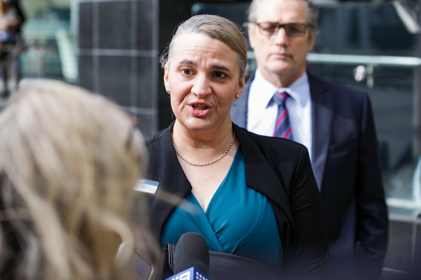 A woman in a blue dress and black jacket speaks to a journalist with a man in a suit standing behind her.