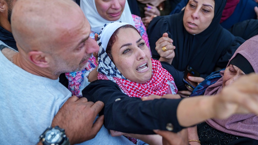 A woman wearing a red checkered keffiyeh cries while supported by a crowd of mourners.