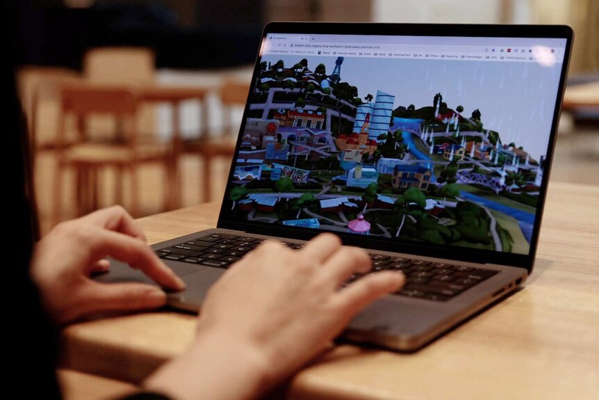 Looking over the shoulder of a girl playing a game on a laptop. A digital illustration of a mountaintop town is on her screen.