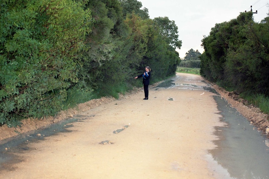 A woman in a black outfit stands pointing on a deserted road with bushland on either side.