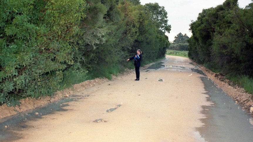 A woman in a black outfit stands pointing on a deserted road with bushland on either side.