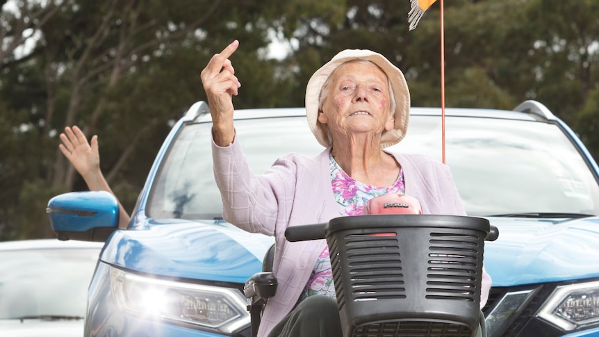 An elderly woman holds up a line of cars behind her mobility scooter and sticks up her middle finger