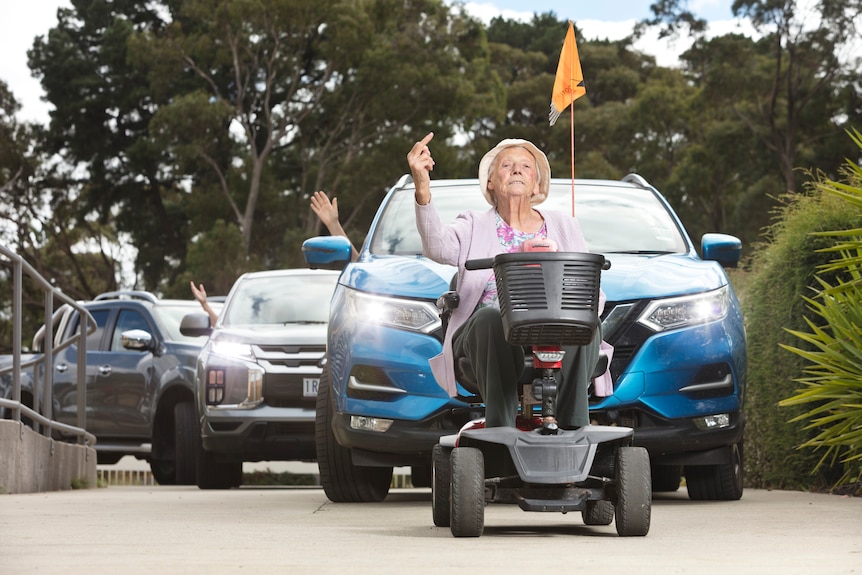 An elderly woman holds up a line of cars behind her mobility scooter and sticks up her middle finger