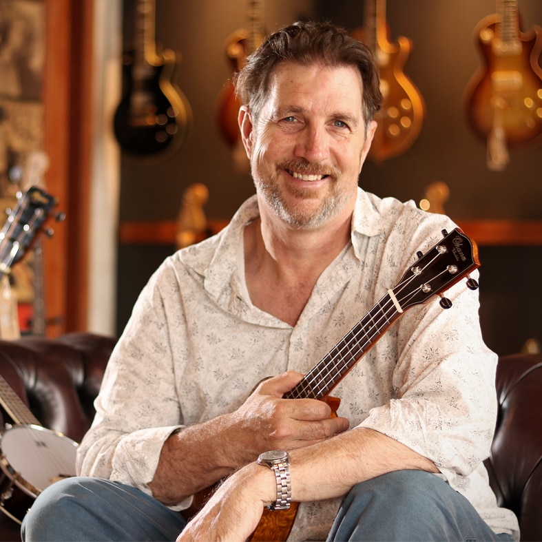 A man with brown hair and a beard, sitting on a couch in a music shop holding a guitar.