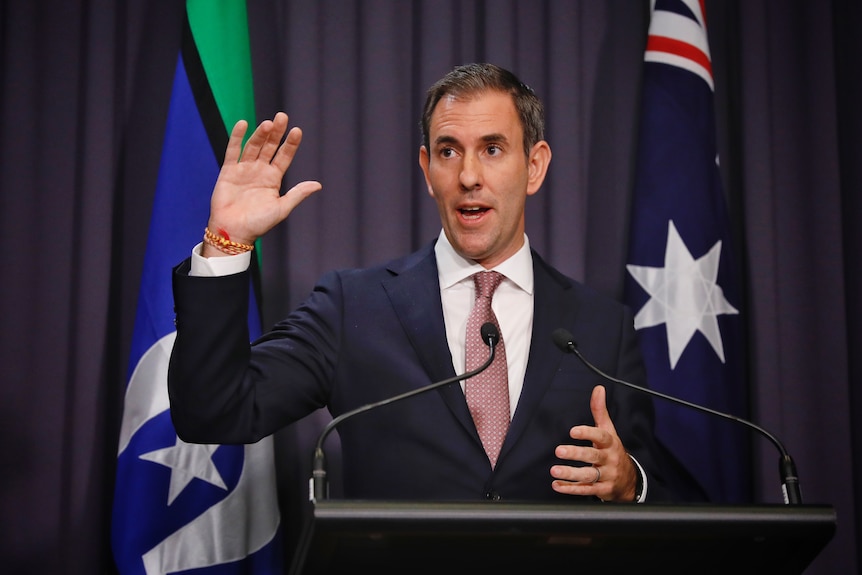 Chalmers raises a hand with a decorative bracelet on his wrist as he speaks in front of a lectern.