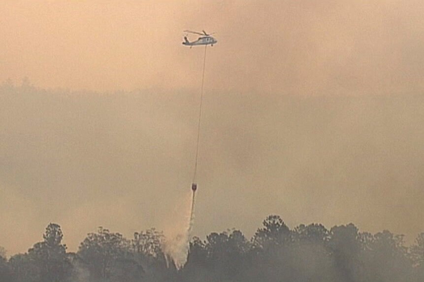 A water-bombing helicopter flying through thick smoke drops water on a fire.