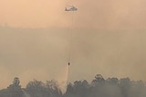 A water-bombing helicopter flying through thick smoke drops water on a fire.
