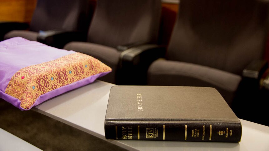 An embriodered purple book-shaped bag next to a black bible sit on a small ledge, rows of chairs in background.