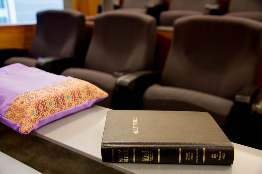 An embriodered purple book-shaped bag next to a black bible sit on a small ledge, rows of chairs in background.