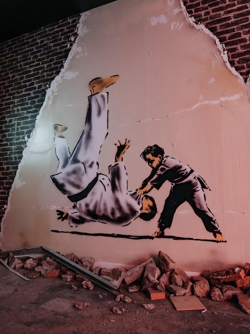 A reproduction of a Banksy mural on a brick wall, showing a karate student throwing his teacher to the ground.