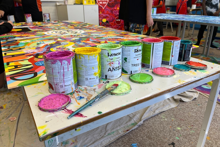 Seven open pain cans sit on a school table in front of a colourful mural.