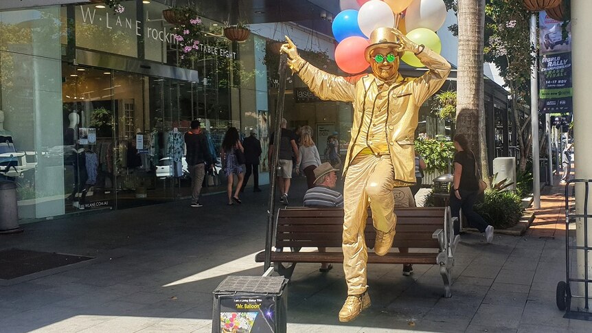 A living statue busker painted in gold is posing for the camera. His illusion makes it seem as if balloons are lifting him.