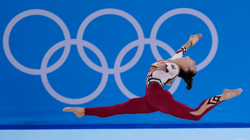 Pauline Schaefer-Betz in a mid-air split wearing the German team red unitard, in front of a banner with Olympic rings. 