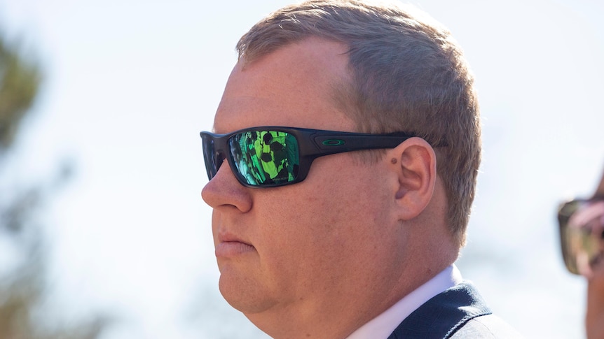 A man looks straight ahead with green tinted sunglasses on