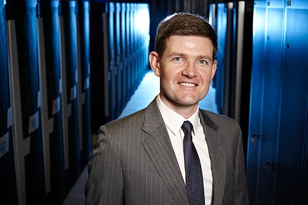 A man in a suit stands in a blue-lit server room.