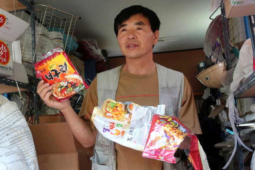 Lee Min Bok holds empty food wrappers, stained labels showing noodles and cookies.