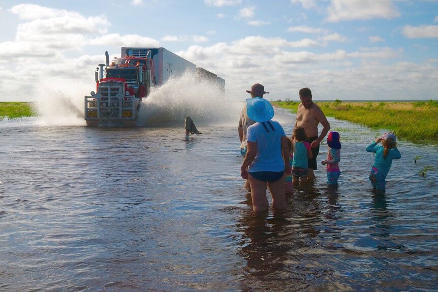 A family stands in knee deep water on Great Northern Highway as a semi trailer drives on the flooded road.