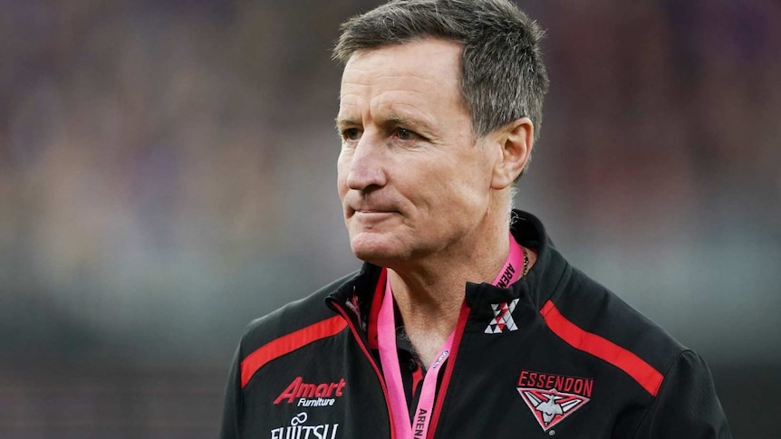 A stern looking middle aged man stands on an AFL match field