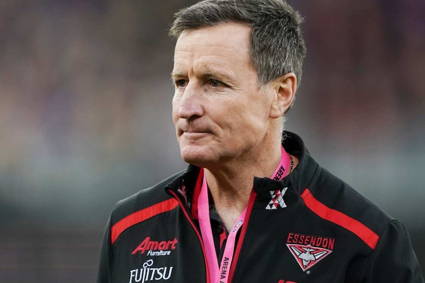 A stern looking middle aged man stands on an AFL match field