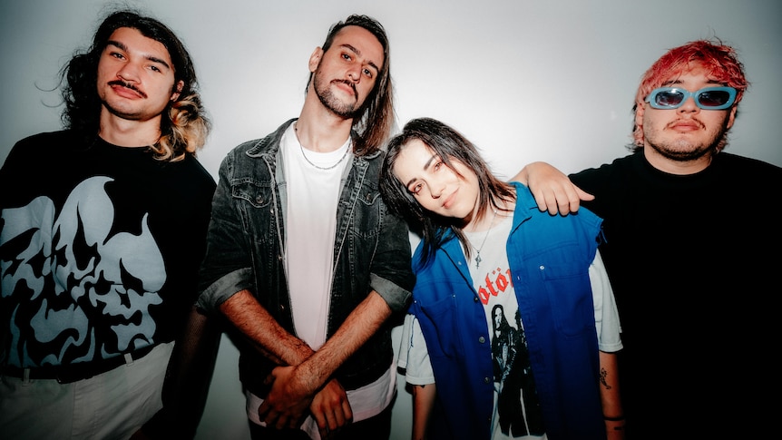 Four people stand against a white wall in black shirts, a denim jacket and a blue cutoff jacket