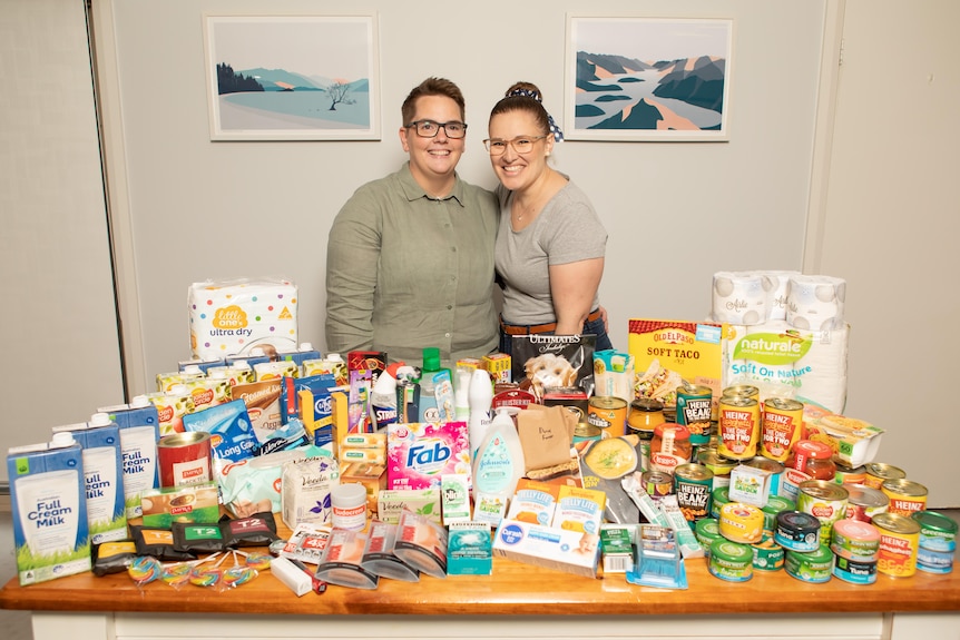 Two women stand behind a table full of food and long life pantry products.