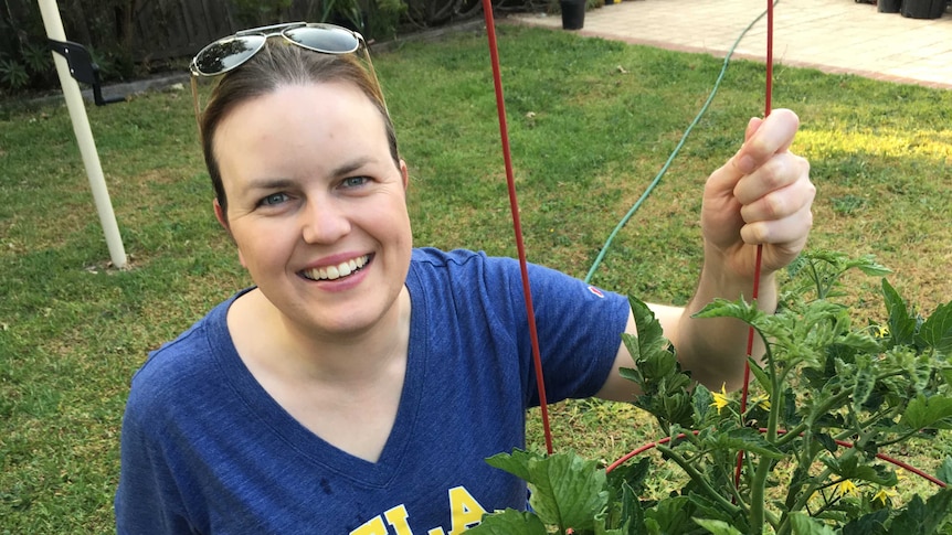 A woman tends to her vegetable patch in her backyard. She's wearing a blue shirt and smiling.