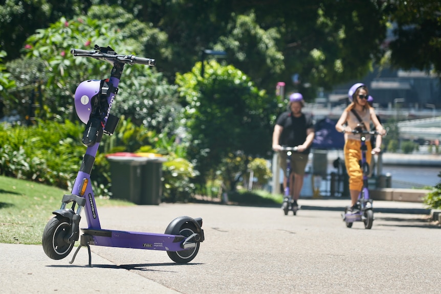 Purple e-scooter parked on footpath with two other people riding on others.