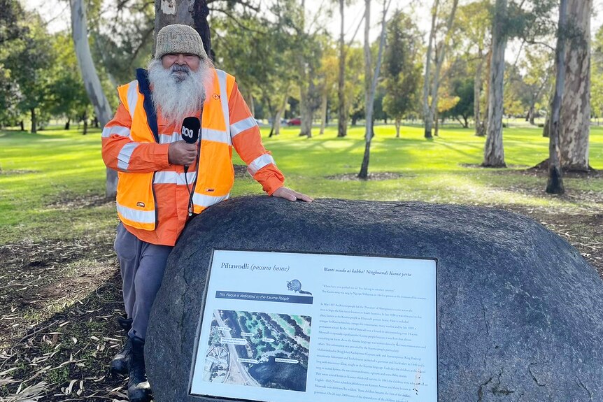Indigenous man in high vis clothes stands by plaque on a rock